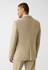 Blazer beige con texture liscia, chiusura a un bottone, rever con incisione e tasche laterali. Include vestibilità sagomata e dettagli di cuciture sottili.