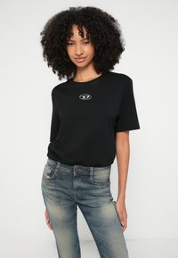 Tricou negru din bumbac cu mâneci scurte, având un logo circular argintiu pe piept, asortat cu blugi din denim albastru decolorați.