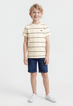 Un garçon blond souriant porte un t-shirt crème à fines rayures noires, un short en denim bleu foncé et des baskets blanches sur un fond blanc.