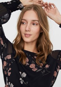 Blouse florale en bleu marine foncé avec des motifs de fleurs roses claires et grises. Manches transparentes et col rond. Tissu doux et texturé avec une coupe décontractée.