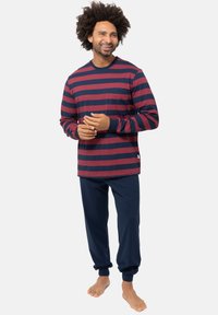 Uomo sorridente in piedi a piedi nudi, con una camicia a maniche lunghe a righe blu e rosse e pantaloni jogging blu, su uno sfondo bianco semplice.