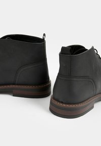 Schwarze Lederstiefel mit abgerundetem Zehenbereich, niedrigem Absatz, gepolstertem Kragen und Schnürdetail. Die Sohle ist mit einem kontrastierenden braunen Akzent versehen.