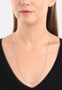 Kobieta nosi cienki srebrny naszyjnik na czarnej bluzce z dekoltem w serek, eksponując szyję i dolną część twarzy na jednolitym tle.