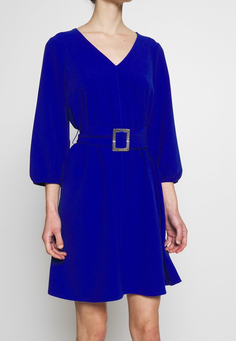 Blå klänning med lång ärm och V-ringning, åtdragen midja och silverfärgad fyrkantig spänne. Släta tyger med utsvängd kjol.