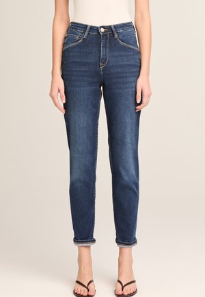 Frau trägt dunkelblaue, hoch taillierte, eng anliegende Jeans mit Umschlag und dunkelbraune Zehensandalen, steht vor einem einfarbigen Hintergrund.