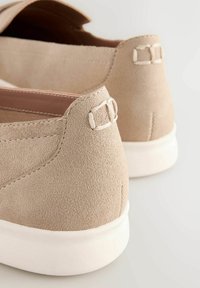 Beige suede slip-on shoes with a white, thick sole. Rear features a decorative stitched accent and a small loop detail near the heel.