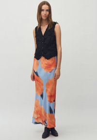 Zwarte textuurvest met knoopsluiting over een blauwe maxirok met bloemenpatroon en oranje bloemen, afgewerkt met een gladde stof. Zwarte enkellaarzen.
