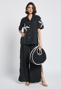 Camicetta nera a maniche corte con ricami floreali, abbinata a pantaloni neri larghi con spacchi laterali. Borsa rotonda nera con bordo beige.