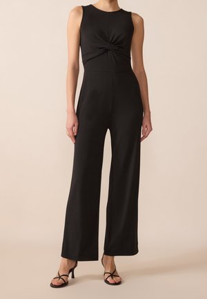 Combinaison noire sans manches avec un détail torsadé à la taille, pantalon large, portée avec des sandales à talons noires à brides.