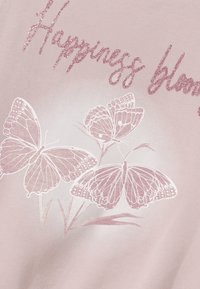 Vaaleanpunainen paita, jossa on valkoisilla ääriviivoilla piirrettyjä kuvioita kahdesta perhosesta yksityiskohtaisine kuvioineen ja kaunokirjoituksella teksti "Happiness blooms".