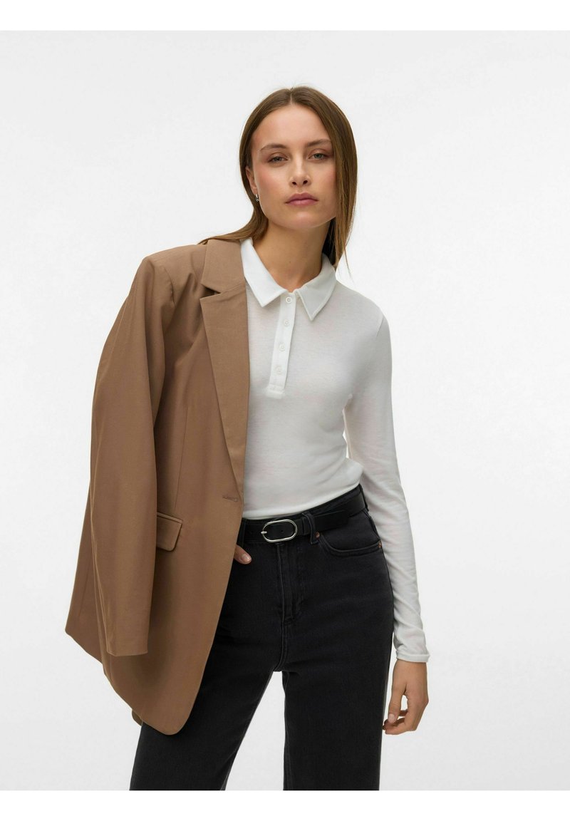 Kobieta ubrana w białą koszulę polo z długim rękawem, czarne jeansy z wysokim stanem i paskiem oraz brązową marynarkę zarzuconą na ramię na białym tle.