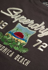 Szary sweatshirt z haftowanym napisem "Superdry 1972" oraz kolorową naszywką przedstawiającą zachód słońca, palmy i zieleń.