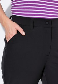 Zwarte broek met een gladde textuur, met een standaard tailleband, een voorkPocket en een zwart knoopsluiting, weergegeven met een hand in de zak.