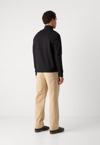 Giacca nera con zip e colletto alto, abbinata a pantaloni beige e scarpe scure, vista da dietro, su uno sfondo bianco.