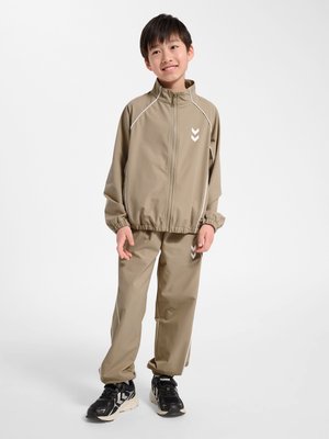 Ung dreng iført beige træningsdragt med lynlås og sorte sneakers, stående og smilende mod en ensartet hvid baggrund.