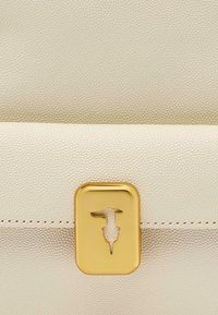 Bolso de cuero texturizado color crema con un cierre dorado que presenta un diseño de logo distintivo. Patrón de grano suave y elevado en toda su superficie.