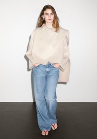 Haut en laine beige oversized avec un détail de design angulaire, associé à un jean large bleu clair et des sandales noires avec des bretelles rouges.