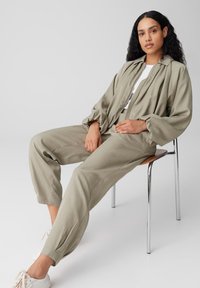 Frau mit langen lockigen Haaren sitzt auf einem Stuhl, trägt eine lockere beige Jacke und Hose, ein weißes Oberteil und weiße Turnschuhe vor einem einfarbigen Hintergrund.