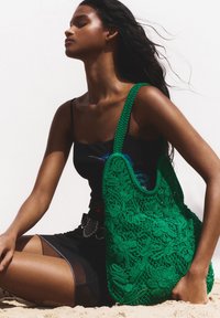 Grön virkad väska med blommönster, strukturerad yta och en bred rem; modellen bär en åtsittande svart topp och en lager-på-lager svart kjol.