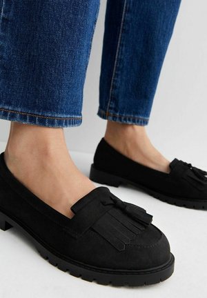 Sorte mokkasiner i ruskind med duske, båret med blå denimjeans på et hvidt gulv, der viser underben og fødder.
