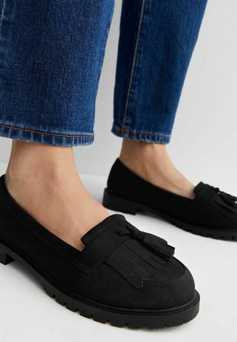 Mocassins en daim noir avec des pompons portés avec un jean denim bleu sur un sol blanc, montrant les jambes et les pieds inférieurs.