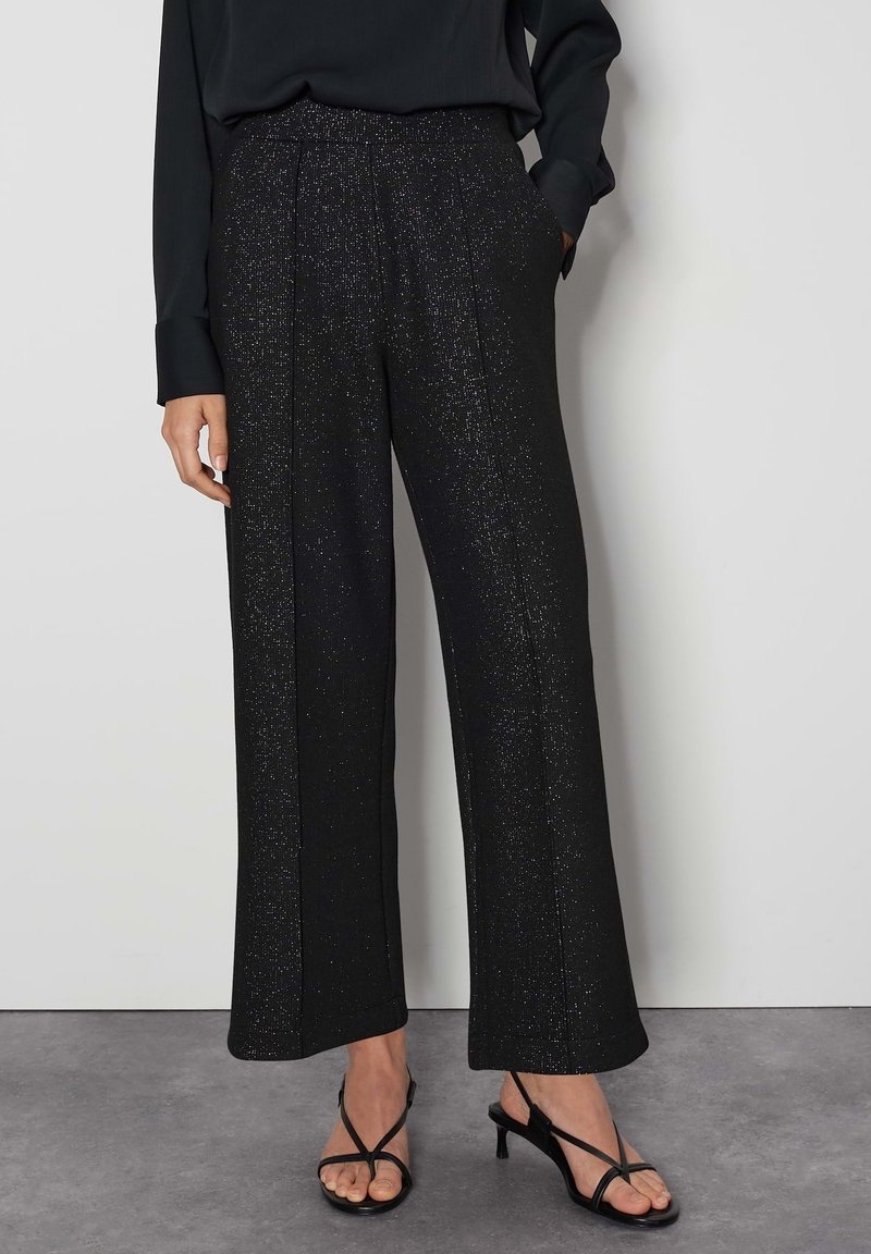 Svarta vida byxor med subtila glittrande detaljer, bärs med svarta klacksandaler med remmar och en inskjuten svart långärmad topp.