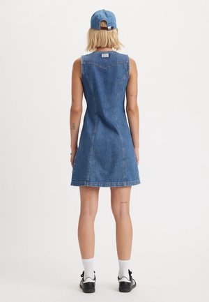 Femme portant une robe en jean bleue sans manches, une casquette de baseball bleue à l'envers, des chaussettes blanches et des baskets noires, vue de dos sur fond blanc.