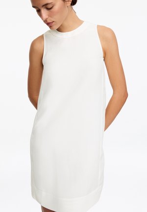 Femme portant une robe blanche sans manches à col montant, debout devant un fond blanc uni.