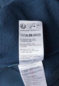 Ciemnoniebieska etykieta z materiału zawierająca instrukcje dotyczące pielęgnacji oraz szczegóły materiałowe w wielu językach. Materiał: 100% poliester, miękka tekstura.