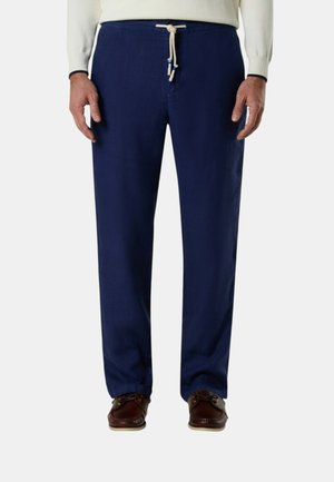 Uomo che indossa pantaloni blu navy con coulisse e scarpe da barca in pelle marrone, in piedi con le mani rilassate ai lati.