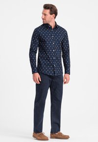 Marineblauw overhemd met kleine, multiculturele patronen, lange mouwen en een standaard kraag, gecombineerd met donkere denim jeans en moccasinschoenen.