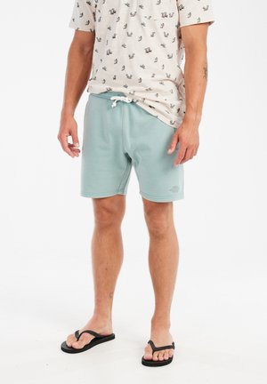 Lichtblauwe sportshorts met een elastische tailleband en trekkoord. Gecombineerd met een crèmekleurig shirt met een klein patroon. Zwarte flip-flops.