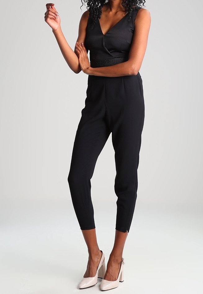 Femme portant une combinaison noire sans manches et des chaussures à talons beiges, debout les bras croisés devant un fond uni.