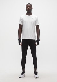 Αθλητικός άνδρας στέκεται, φορώντας λευκό T-shirt της Puma, μαύρα κολάν, μαύρα γάντια χωρίς δάχτυλα και λευκά αθλητικά παπούτσια, σε λευκό φόντο.