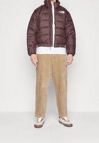 Brązowa kurtka puchowa z wysokim kołnierzem i pikowanym wzorem, noszona na białym t-shircie. W zestawieniu z jasnobrązowymi spodniami z korduroy i trampkami.