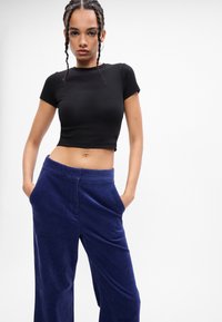 Νέος/νέα με πλεγμένα μαλλιά που φοράει ένα εφαρμοστό μαύρο crop top και χαλαρές σκούρες μπλε παντελόνες από κοτλέ, με τα χέρια στις τσέπες.