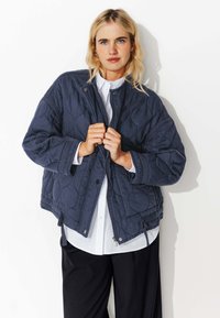 Veste matelassée bleu marine avec un zip sur le devant, un col montant et une taille élastiquée. Tissu texturé avec une finition lisse, dotée de cordons de serrage sur les côtés.