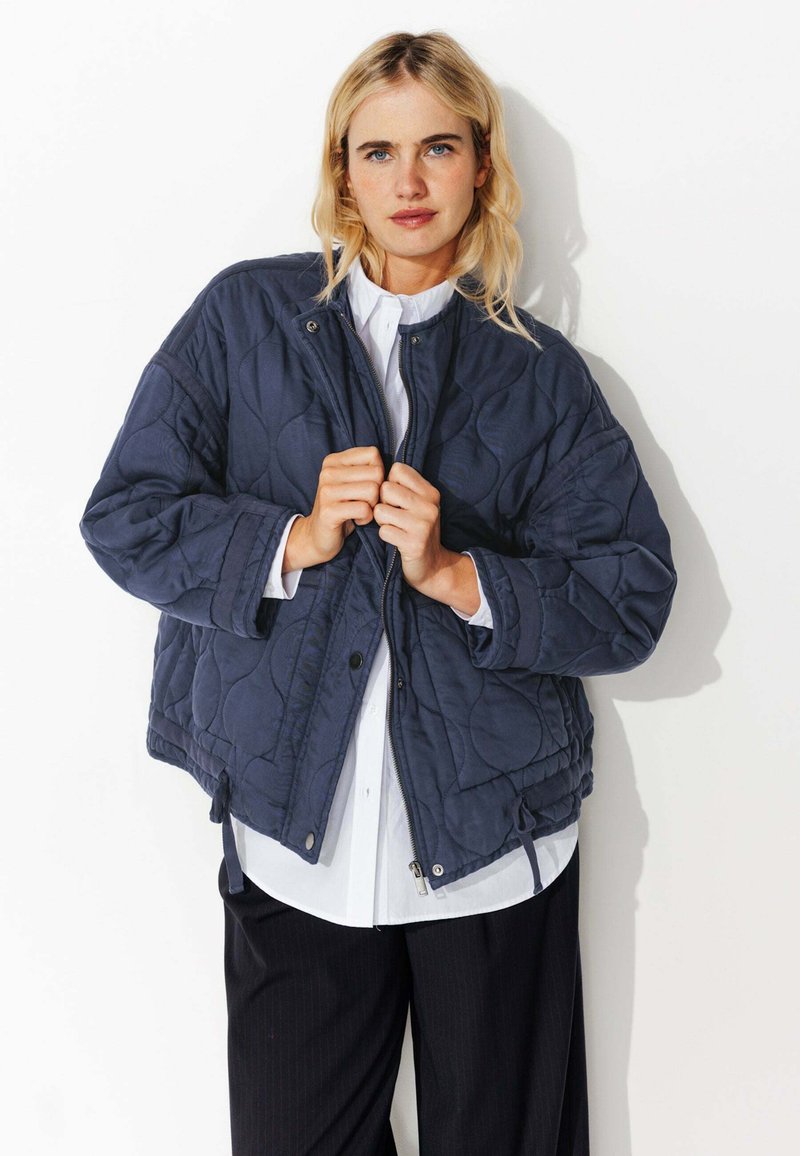 Veste matelassée bleu marine avec un zip sur le devant, un col montant et une taille élastiquée. Tissu texturé avec une finition lisse, dotée de cordons de serrage sur les côtés.