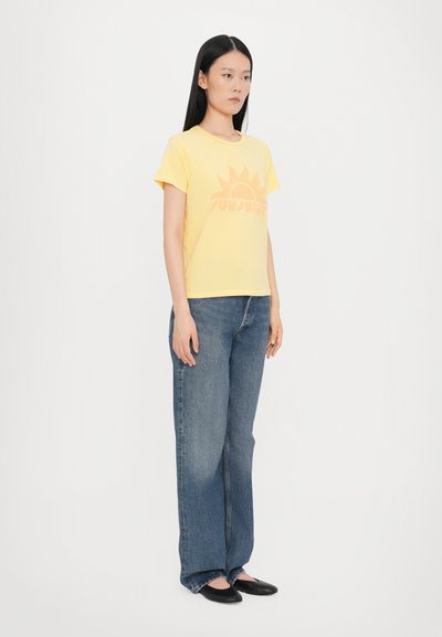 Gul bomull T-skjorte med en solgrafikk i oransje. Kombinert med lys blå denimjeans. Modellen står og har på seg svarte leiligheter.