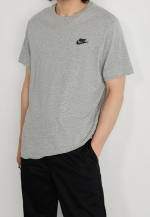 Person trägt ein hellgraues Nike-T-Shirt mit schwarzem Logo auf der Brust und schwarze Hose, eine Hand in der Tasche.