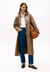 Lungo trench coat beige con chiusure a bottoni, indossato sopra una camicia bianca e jeans blu. Una borsa in pelle marrone e mocassini neri completano l'outfit.