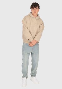 Un jeune homme portant un sweat à capuche beige oversize, un jean bleu clair et des baskets blanches, se tenant devant un fond gris clair uni.