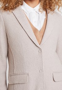 Donna che indossa un cappotto texturizzato beige chiaro sopra una camicia bianca con colletto e un gilet a V color cammello.