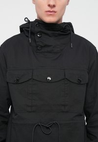 Black jacket made of fabric with a high collar, featuring large front pockets, black buttons, and an adjustable drawstring at the waist.