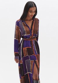Abito lungo con bottoni di colore viola, decorato con motivi geometrici in marrone e arancione. Presenta una vita con cintura e uno spacco laterale. Tessuto dalla texture liscia.
