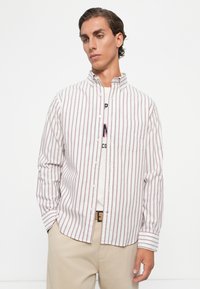 Young man wearing a white and red vertical striped button-up shirt over a graphic t-shirt, with beige pants, standing against a white background.