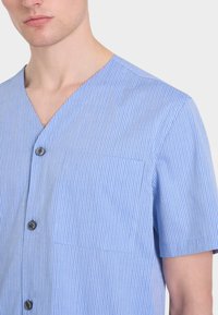 Camicia a maniche corte di colore azzurro chiaro con sottili righe verticali, scollo a V, bottoni e una singola tasca sul petto. Texture liscia.