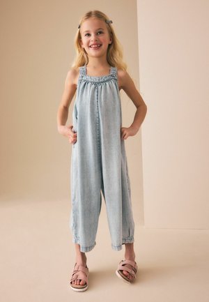 Lächendes junges Mädchen mit langen blonden Haaren, das einen hellen, ärmellosen Jeans-Jumpsuit und pinke Sandalen trägt und mit den Händen in den Taschen steht.