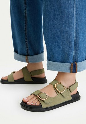 Füße, die hochgekrempelte blaue Jeans und olivgrüne Sandalen mit goldenen Schnallen auf schwarzen Sohlen vor weißem Hintergrund tragen.