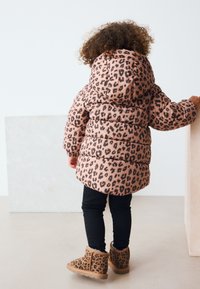 Jachetă matlasată cu imprimeu leopard roz, cu glugă, colanți negri și cizme maro cu imprimeu leopard. Jacheta are secțiuni matlasate și mansete elastice.